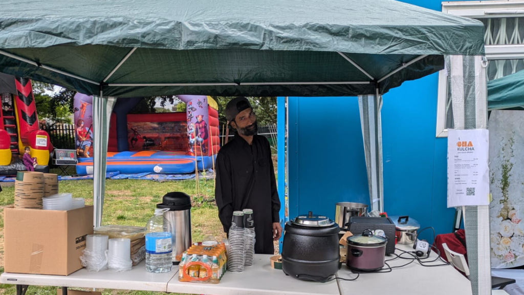 Faiq is standing at the pop up stall. In front is layed out various paraphenalie for selling food. A gazebo covers the whole stall.