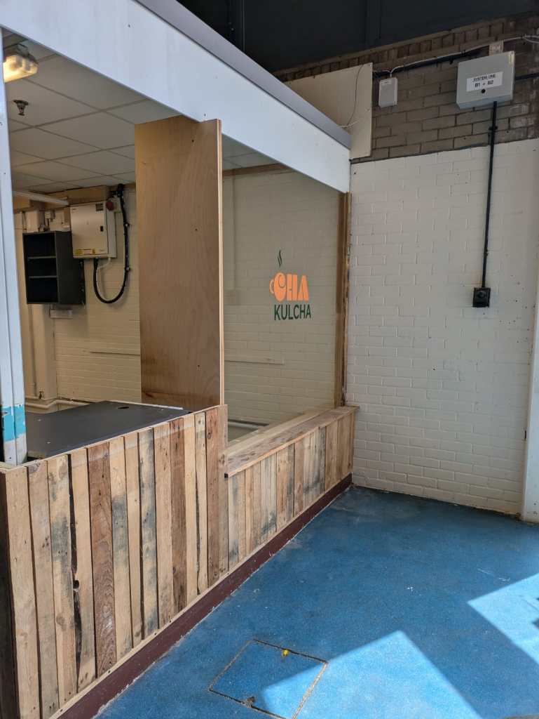 A freshly painted white brick wall in the background. Coming out from that wall is on of the counters to our stall. The is a glass wall with a "Cha Kulcha" decal. 